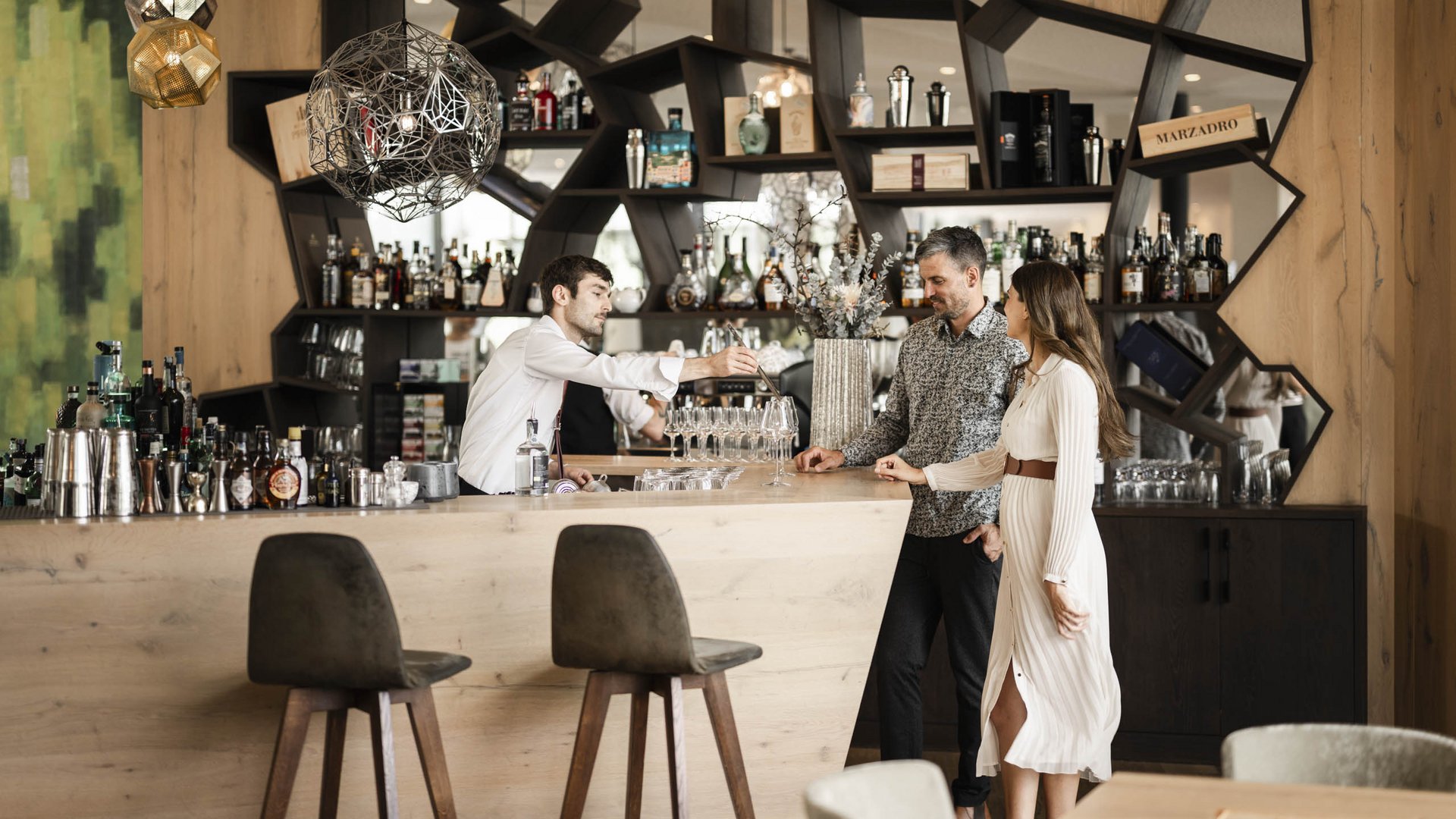 Bar mit Barkeeper und zwei Gästen vor einem modernen Holzregal voller Flaschen
