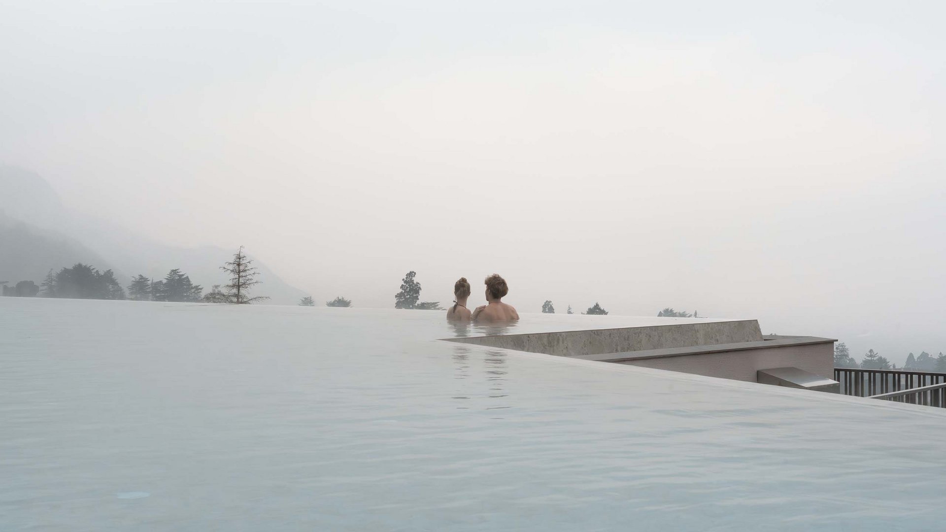 Schenna Resort Zwei Personen entspannen im Infinity-Pool bei Nebel und bewölktem Himmel.